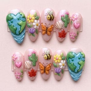 May include: A set of ten artificial nails with a light pink base, each adorned with 3D designs. The designs include a globe, flowers, bees, butterflies, cacti, seashells, maple leaves, and hearts. The nails are arranged in two rows of five.