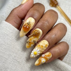 Puede incluir: Uñas almendradas amarillas con diseños 3D de miel y abejas. Una uña presenta un tarro de miel con una abeja, otra tiene un cucharón de miel y miel goteando con abejas, y otra tiene un patrón de panal con una margarita.