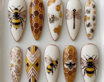 Puede incluir: Un juego de diez uñas postizas en forma de almendra con diseños intrincados en blanco, amarillo, marrón y oro metálico. Los patrones incluyen abejas detalladas, estructuras de panal, una libélula y motivos botánicos con bordes dorados.