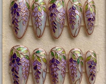 May include: A set of ten almond-shaped press-on nails featuring a pearlescent white base with hand-painted wisteria floral designs in shades of purple and green, outlined with thin metallic gold lines in a stained-glass style.