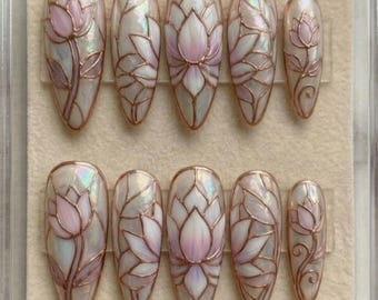 May include: Set of ten oval-shaped press-on nails. Each nail features a pearlescent base with a pink and white lotus flower design outlined in a copper-colored metal. The lotus flowers are in various stages of bloom.