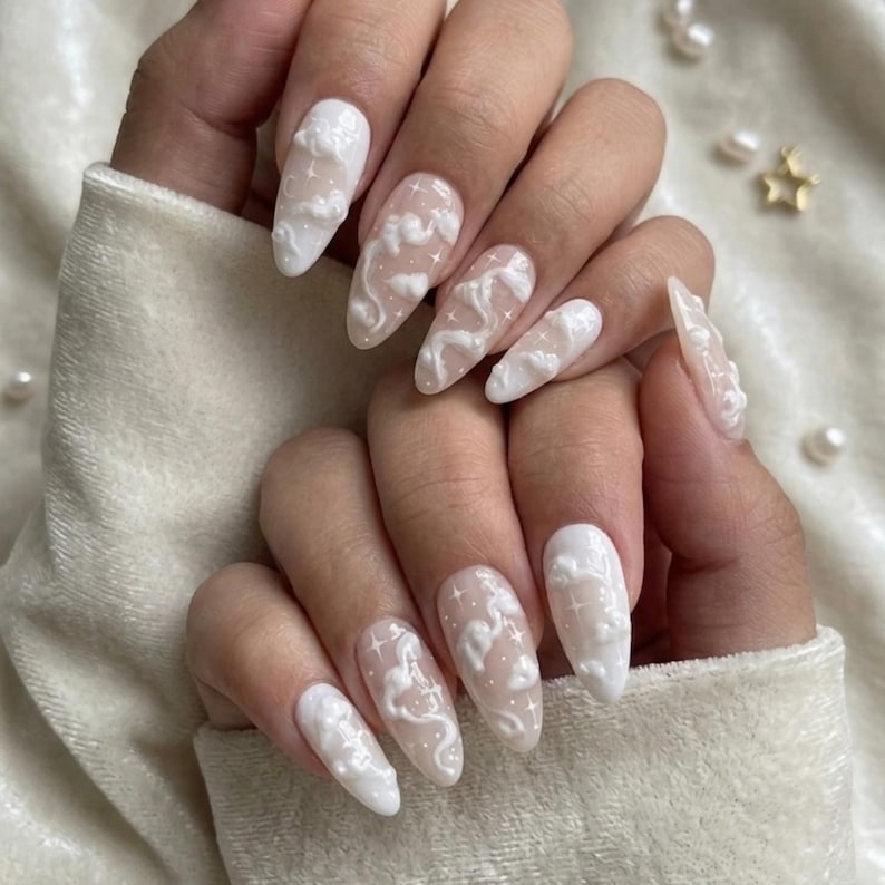 May include: Almond-shaped, white acrylic nails with a cloud design. The nails feature 3D cloud embellishments and small star accents. The design is set against a neutral background, creating a dreamy aesthetic.