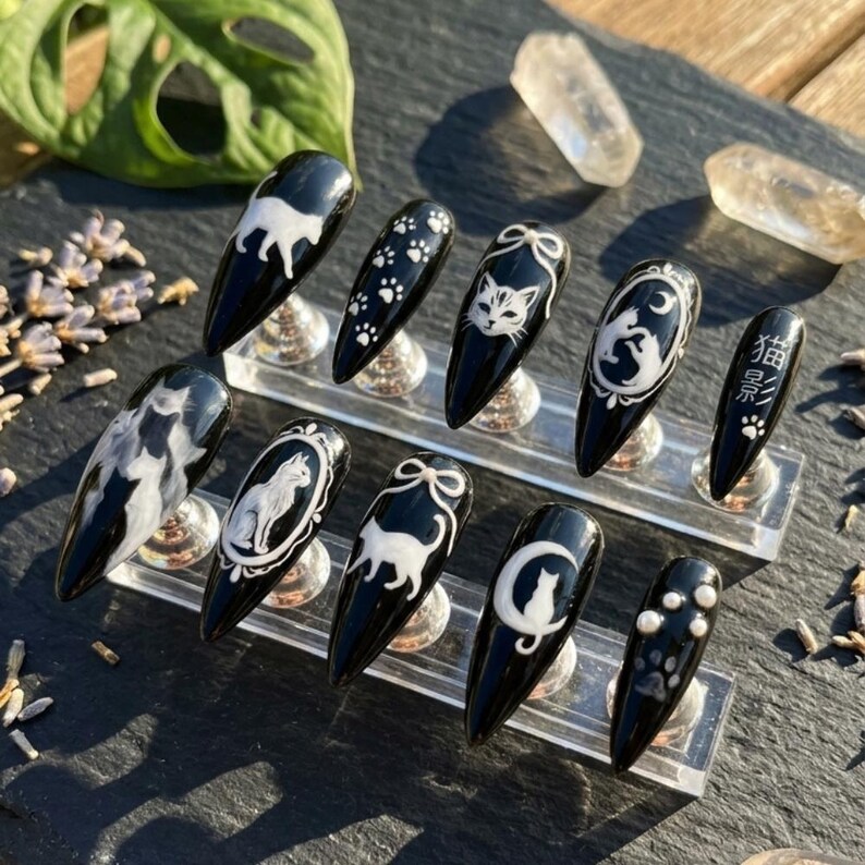 May include: Black nail set with white cat designs, paw prints, and Japanese text. The nails are almond-shaped and displayed on a clear stand. The designs include cat silhouettes, cat faces, and a cat with a moon.