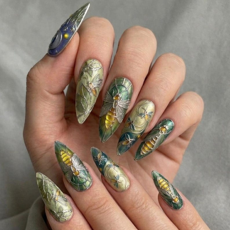 May include: A hand showing long, almond-shaped nails with detailed designs. The nails are painted in green, gold, and blue, with insect and floral patterns. One nail has a celestial design with a moon and stars.