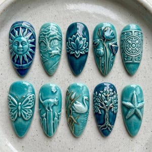 May include: Ten teardrop-shaped ceramic tiles in shades of teal and blue with a crackle glaze finish. Each tile features a raised relief design including a sun, a tree with a rabbit, a lotus flower, poppies, a geometric pattern, a butterfly, a cat, a swan, a tree, and a starfish.