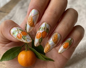 May include: Oval-shaped nail art featuring a citrus fruit design. The nails have a pearlescent base with orange fruit and green leaf accents, outlined in gold. A fresh orange and leaves are held in the hand.
