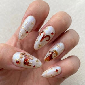 Puede incluir: Una mano muestra uñas largas y ovaladas con un acabado blanco nacarado. Cada uña presenta detalles dorados, gemas rojas y símbolos del zodiaco Aries en rojo y marrón. El diseño incluye estrellas y patrones de constelaciones.