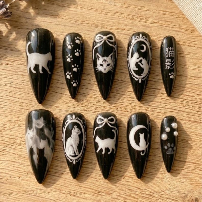 May include: Set of ten black press-on nails with white cat-themed designs. The designs include cat silhouettes, paw prints, and the Japanese text "Cat Shadow". Each nail has a glossy finish and a pointed shape.