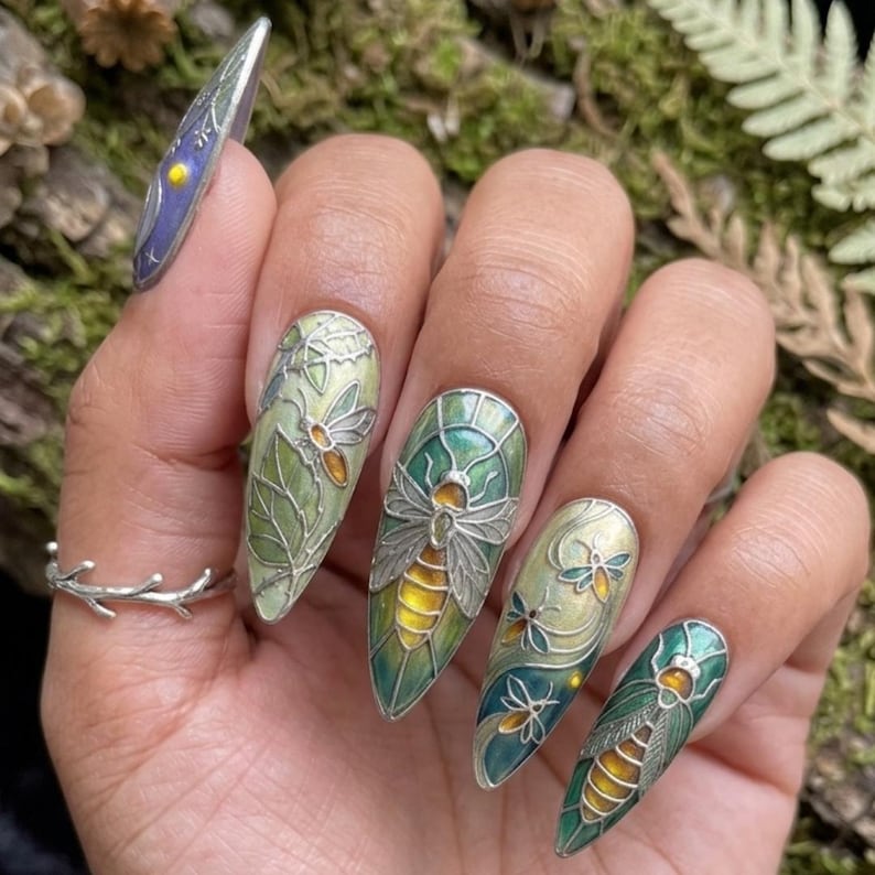 May include: A hand showing long, pointed fingernails with detailed designs. The nails have a silver insect and leaf pattern, with green, yellow, and silver highlights. One nail features a blue and yellow celestial design. A silver twig ring is also visible.