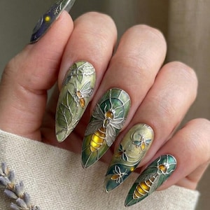 May include: Intricate nail art with a nature theme. The nails are painted in green and yellow hues, adorned with detailed silver insect and leaf designs. The design incorporates various insect and leaf patterns.