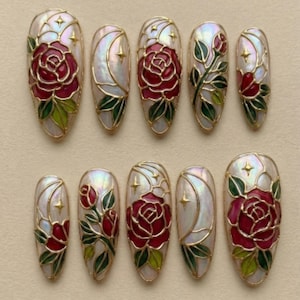 Puede incluir: Diez uñas postizas ovaladas con acabado nacarado y contorno dorado. Cada uña presenta un diseño de rosa roja con hojas verdes, una luna creciente y estrellas doradas.