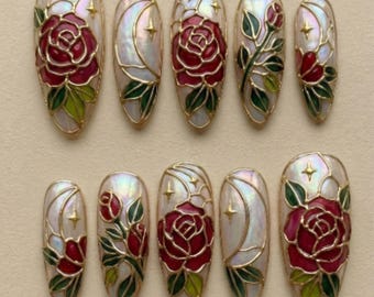 May include: Set of ten artificial nails with a pearlescent finish, featuring a gold outline. Each nail showcases a red rose design with green leaves, a crescent moon, and gold stars. The nails are oval-shaped.