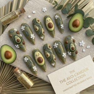 Pode incluir: Um conjunto de unhas reutiliz&aacute;veis em forma de am&ecirc;ndoa com tema de abacate. As unhas s&atilde;o verde-oliva com desenhos de abacate, flores brancas e detalhes de glitter dourado. O texto no cart&atilde;o diz "The Avo-L'Amour Collection, Handcrafted Reusable Nails."