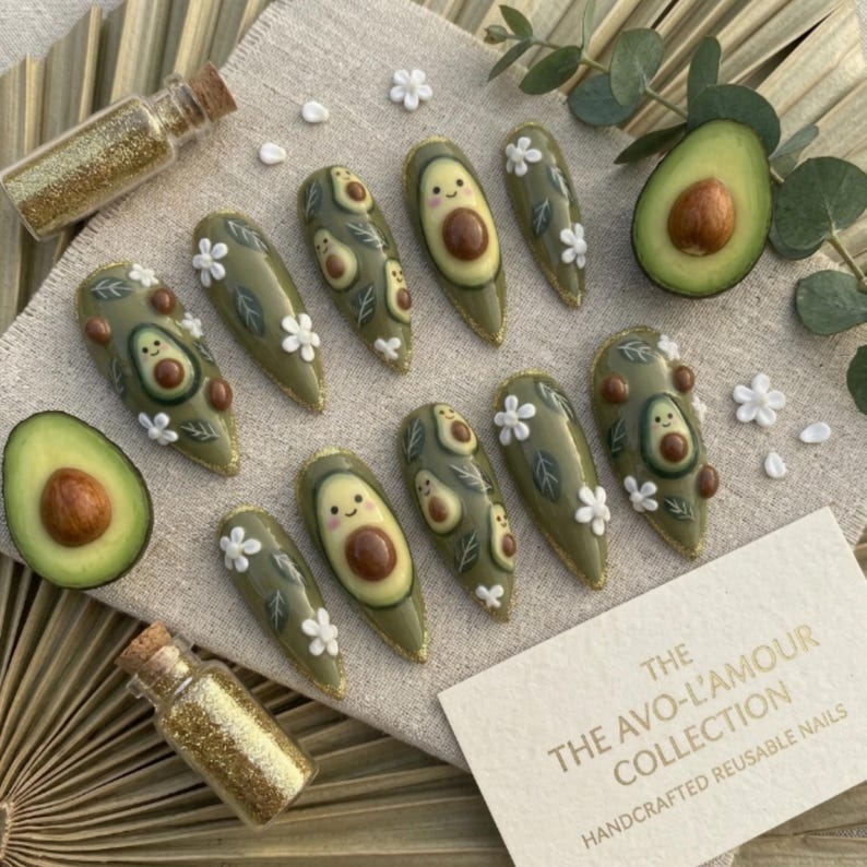 Pode incluir: Um conjunto de unhas reutiliz&aacute;veis em forma de am&ecirc;ndoa com tema de abacate. As unhas s&atilde;o verde-oliva com desenhos de abacate, flores brancas e detalhes de glitter dourado. O texto no cart&atilde;o diz "The Avo-L'Amour Collection, Handcrafted Reusable Nails."