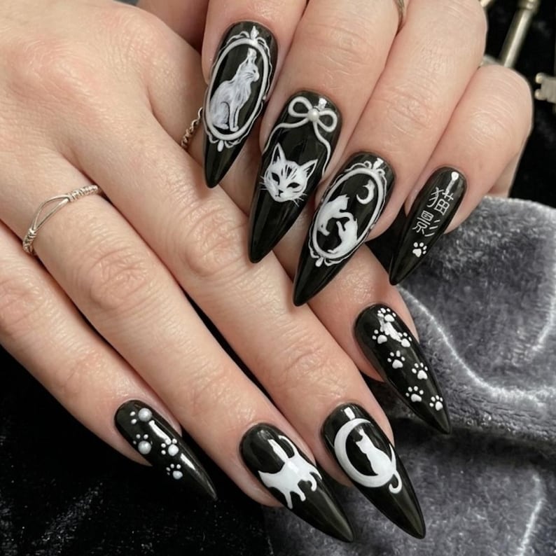 May include: Black stiletto nails with white cat-themed designs. Each nail features a different cat illustration, including a cat in a frame, a cat face, and paw prints. The nails also have Japanese text.