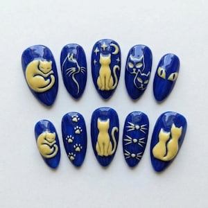 May include: A set of ten almond-shaped press-on nails featuring a deep blue base color with raised, cream-colored cat-themed designs. The patterns include sleeping cats, cat silhouettes, paw prints, whiskers, and two cats sitting together.