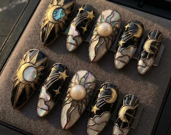Puede incluir: Un conjunto de diez uñas postizas en forma de almendra. Cada uña presenta diseños celestiales, incluyendo soles, lunas, estrellas y nubes, con detalles dorados e iridiscentes. Las uñas son principalmente negras, blancas y doradas.