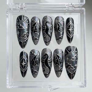 May include: A set of ten press-on nails in a clear plastic case. The nails are oval-shaped with a dark gray and black design. Each nail features a dragon, clouds, and star patterns.