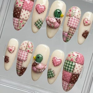 Könnte beinhalten: Ein Set aus zehn künstlichen Nägeln mit cremefarbener Basis. Jeder Nagel hat ein einzigartiges Design, darunter Patchworkmuster in Rosa, Braun und Grün, Blumendrucke und 3D-Enten- und Herzverzierungen. Die Nägel sind für einen einzigartigen und dekorativen Look konzipiert.