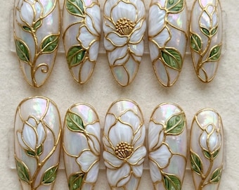 Puede incluir: Un conjunto de diez uñas artificiales de forma ovalada. Cada uña presenta un diseño floral blanco con contornos y detalles dorados. Se incorporan hojas verdes en el diseño, creando una estética delicada y elegante.