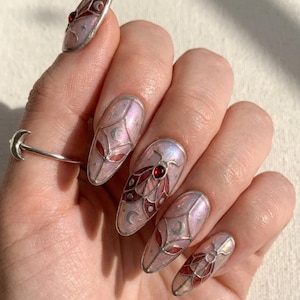 Könnte beinhalten: Nahaufnahme einer Hand mit langen, oval geformten Nägeln. Die Nägel sind mit einem hellrosa schillernden Lack bemalt und weisen aufwendige Designs in Rot, Silber und Weiß auf. Ein silberner Ring mit einem Halbmond befindet sich am Ringfinger.