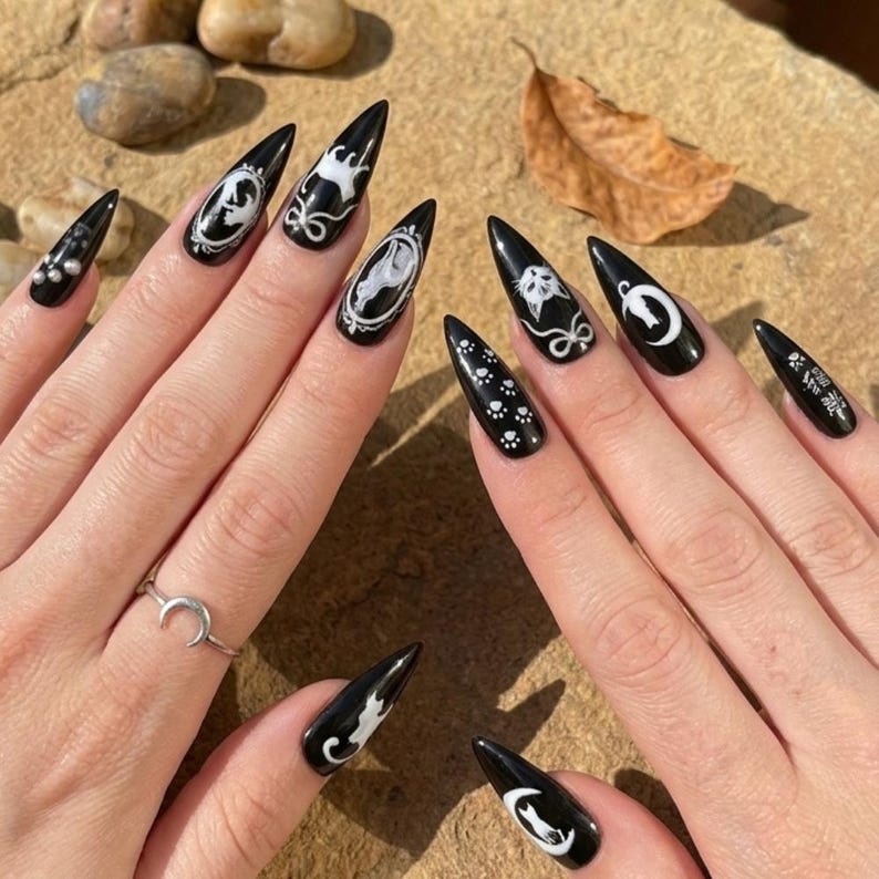 May include: Black stiletto nails with white gothic-themed designs. The nail art includes cat silhouettes, moons, and ornate frames. A silver crescent moon ring is visible on one finger.