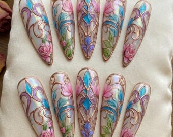 May include: Ten long, almond-shaped artificial nails. Each nail features a stained-glass design with pink roses, blue and purple accents, and a copper-colored outline. The nails are arranged on a cream-colored surface.