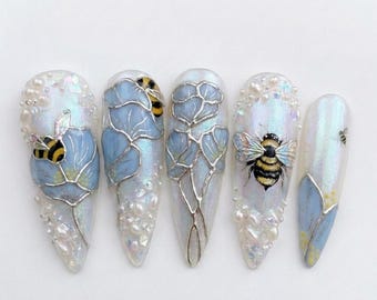 May include: Five long, almond-shaped artificial nails with a pearlescent finish. Each nail features a different design, including blue floral accents, silver outlines, and detailed bee illustrations. Some nails are adorned with small, pearl-like embellishments.