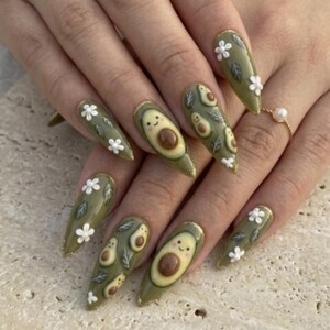 Pode incluir: Unhas artificiais longas em forma de am&ecirc;ndoa com tema de abacate. As unhas s&atilde;o pintadas de verde-oliva com desenhos de abacate, flores brancas e pontas douradas com glitter. Algumas unhas apresentam rostos de abacate sorrindo.