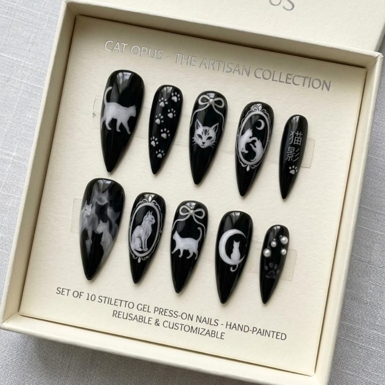 May include: A set of ten black stiletto gel press-on nails with white cat-themed designs. The designs include cat silhouettes, paw prints, and cat illustrations. The text "CAT OPUS - THE ARTISAN COLLECTION" is visible.