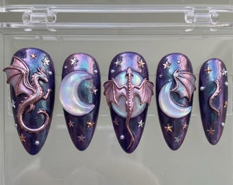 May include: Five purple and blue press-on nails with a fantasy theme. Each nail features a different design, including a dragon, a crescent moon, and stars. The dragon and moon are 3D, with a metallic finish. The nails are in a clear plastic case.
