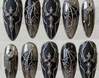 May include: A set of ten press-on nails featuring a pointed almond shape. The designs include black and metallic silver raised motifs of gothic crosses and bat wings, accented with small black rhinestones on a dark, glossy, and translucent base.