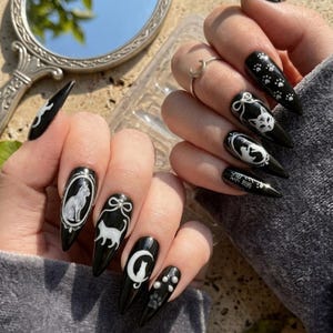 May include: Black stiletto nails with white cat-themed designs. The nails feature various cat illustrations, paw prints, and Japanese text. The designs are set against a glossy black background, creating a striking contrast.