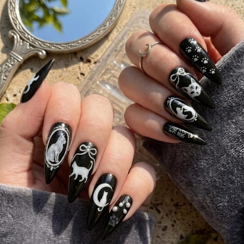 May include: Black stiletto nails with white cat-themed designs. The nails feature various cat illustrations, paw prints, and Japanese text. The designs are set against a glossy black background, creating a striking contrast.
