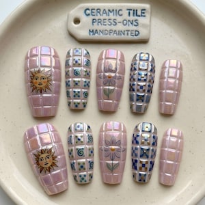 May include: A set of ten press-on nails with a grid-like, iridescent pink finish. Designs include a yellow sun, blue and green dots, a purple flower, and a blue and white checkered pattern. A ceramic tag reads CERAMIC TILE PRESS-ONS HANDPAINTED.