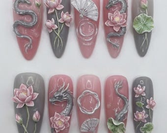 May include: A set of ten artificial nails with a pink and gray color scheme. Each nail features 3D designs, including dragons, lotus flowers, and fans. The nails are oval-shaped and have a glossy finish, showcasing intricate details.