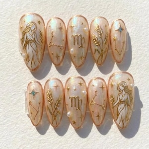 May include: A set of ten almond-shaped artificial nails. Each nail is painted with a light peach base and features gold accents. Designs include zodiac symbols, wheat stalks, stars, and a figure of a woman. Small pearls are also used as embellishments.