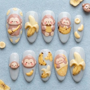 Könnte beinhalten: Zehn hellblaue Kunstnägel mit 3D-Affen- und Bananen-Designs. Die Affen sind braun und beige, die Bananen gelb. Einige Nägel zeigen einen Affen, der eine Banane oder ein Getränk hält.