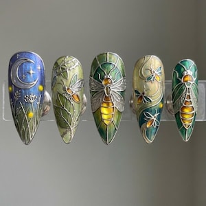 May include: Five intricately designed nail art pieces. Each nail displays a unique design, including a moon and stars, insects, and foliage, with a colour scheme of blues, greens, and yellows. The nails are mounted on a clear display, showcasing the detailed artwork.