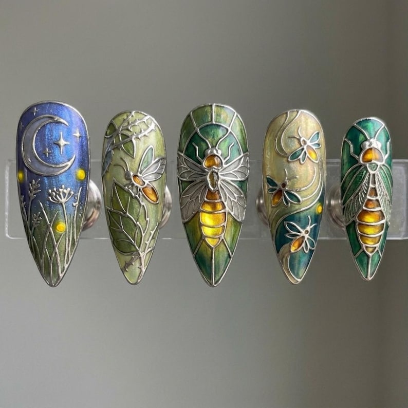 May include: Five intricately designed nail art pieces. Each nail displays a unique design, including a moon and stars, insects, and foliage, with a colour scheme of blues, greens, and yellows. The nails are mounted on a clear display, showcasing the detailed artwork.