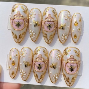 Peut inclure: Ensemble de dix faux ongles ovales. Chaque ongle est peint avec une base blanche nacrée et présente des accents dorés, dont une théière avec une abeille, des motifs floraux et de petites perles. Certains ongles ont un motif de goutte d'or.
