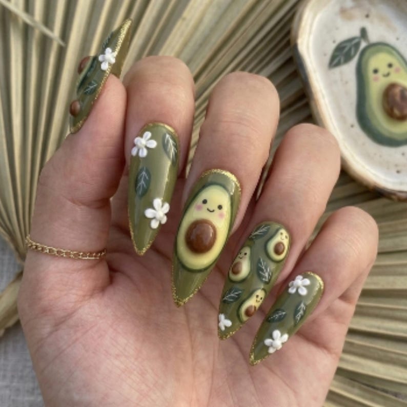 Pode incluir: Unhas em formato am&ecirc;ndoa verde-oliva com desenhos de abacate e florais. As unhas apresentam um abacate sorrindo, pequenas fatias de abacate, flores brancas e detalhes em glitter dourado. Um anel de ouro &eacute; vis&iacute;vel.