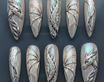 May include: A set of ten iridescent, almond-shaped artificial nails. Each nail features a silver dragon design, with some showcasing dragon heads, wings, or scaled bodies. The nails have a pearlescent finish, reflecting various colors.