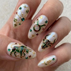 May include: A hand with long, almond-shaped nails, each adorned with a unique design. The designs include gold constellations, zodiac symbols, and a bull, accented with green, red, and gold embellishments. The nails have a pearlescent finish.