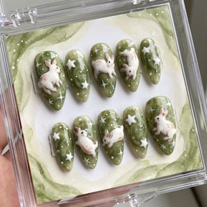 May include: A set of ten artificial nails with a green marble effect, featuring white rabbits and star designs. The nails are oval-shaped and presented in a clear plastic case.