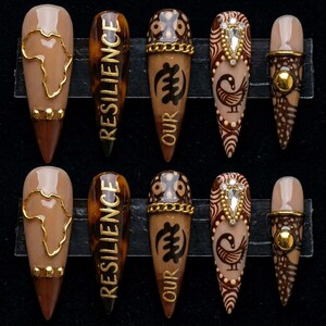 May include: A set of ten long, almond-shaped artificial nails. The nails feature various designs, including a gold outline of Africa, the word "RESILIENCE", and other cultural symbols. Colors include brown, tan, and gold.