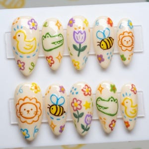 May include: Set of ten press-on nails with a cream-colored base, each adorned with colorful, hand-drawn designs. The designs include a yellow duck, a green alligator, a purple tulip, a bee, a lion, and flowers.