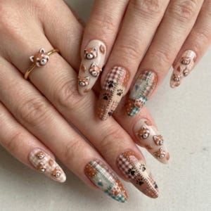 Puede incluir: Uñas almendradas con diversos diseños. Algunas uñas presentan una base beige con caras de panda rojo en 3D, otras tienen patrones de cuadros y huellas de patas. También se ve un anillo dorado con una cara de panda rojo.
