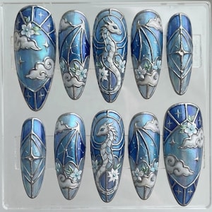 May include: A set of ten almond-shaped press-on nails featuring a blue and silver stained glass design. The artwork includes intricate silver outlines, white dragon figures, clouds, and floral accents set against a shimmering blue gradient background.