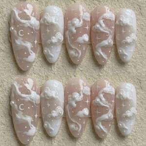 May include: Set of ten almond-shaped artificial nails. Each nail features a light pink base with white cloud and star designs. Some nails have a small letter "C" on them. The nails are arranged in two rows of five.